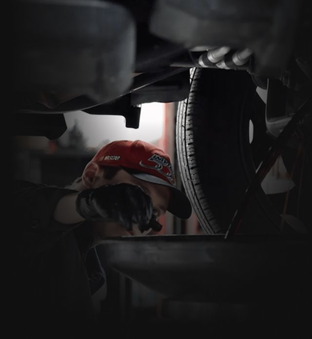 Mechanic draining oil from a car, wearing a red cap, under the vehicle, garage setting. | Auto Medics of Missoula