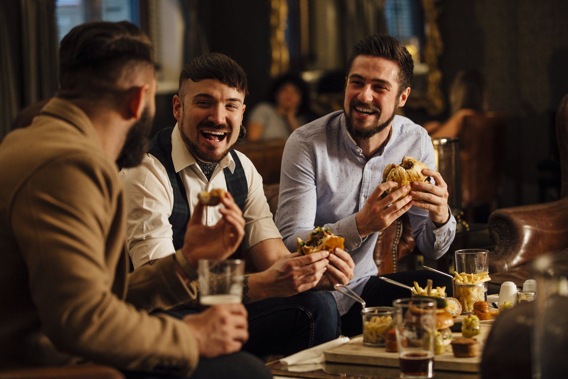 Three men are sitting at a table eating hamburgers and drinking beer.