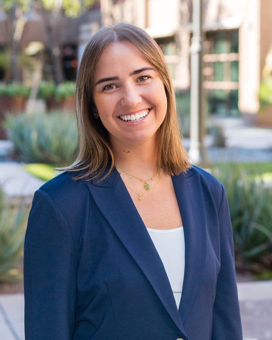 A woman is standing in front of a desert backdrop. Shoulder length brown hair smiling at the camera wearing a blue suit with a white undershirt. 