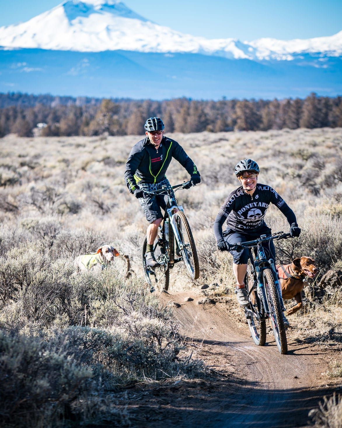 A man and a woman are riding bikes on a trail with a dog.