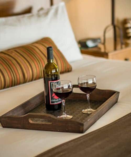 A bottle of wine and two glasses of wine on a wooden tray on a bed