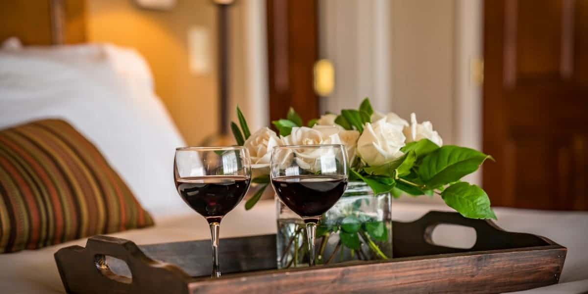 A wooden tray with two glasses of wine and a vase of flowers on a bed.