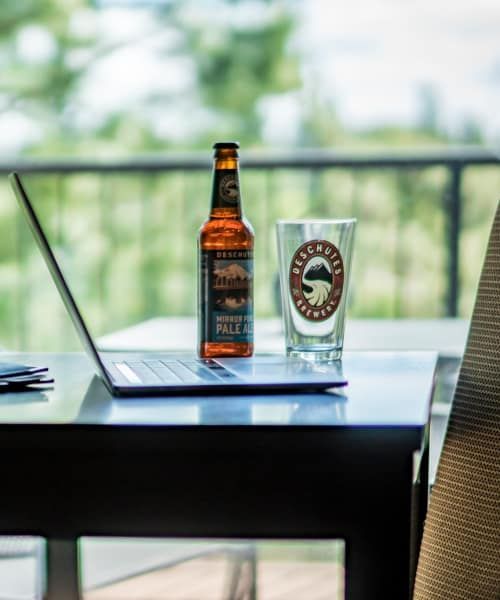 A bottle of beer sits on a table next to a laptop and a glass