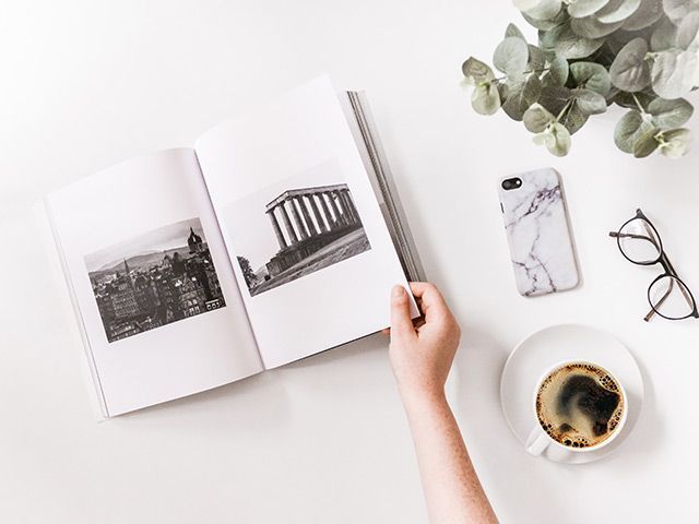 A person is reading a book next to a cup of coffee and a cell phone.