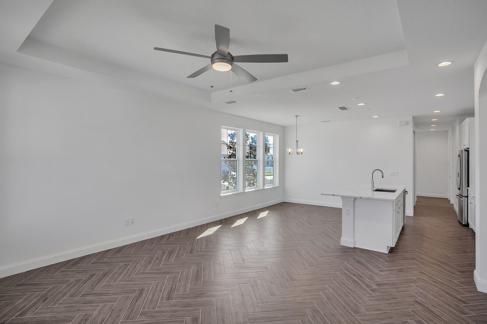 An empty living room with a ceiling fan and a kitchen in the background.