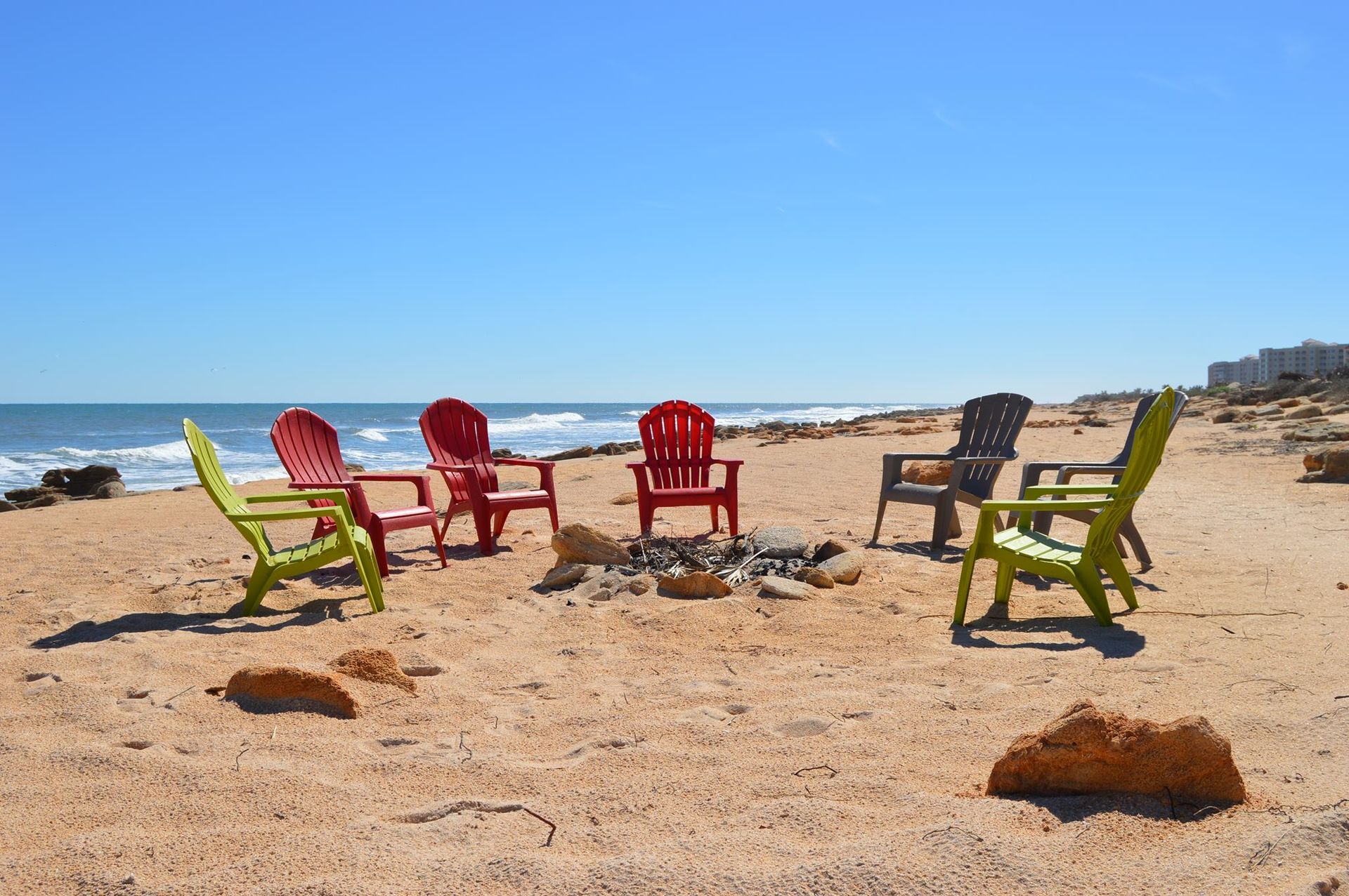 A group of chairs are sitting around a fire pit on the beach.