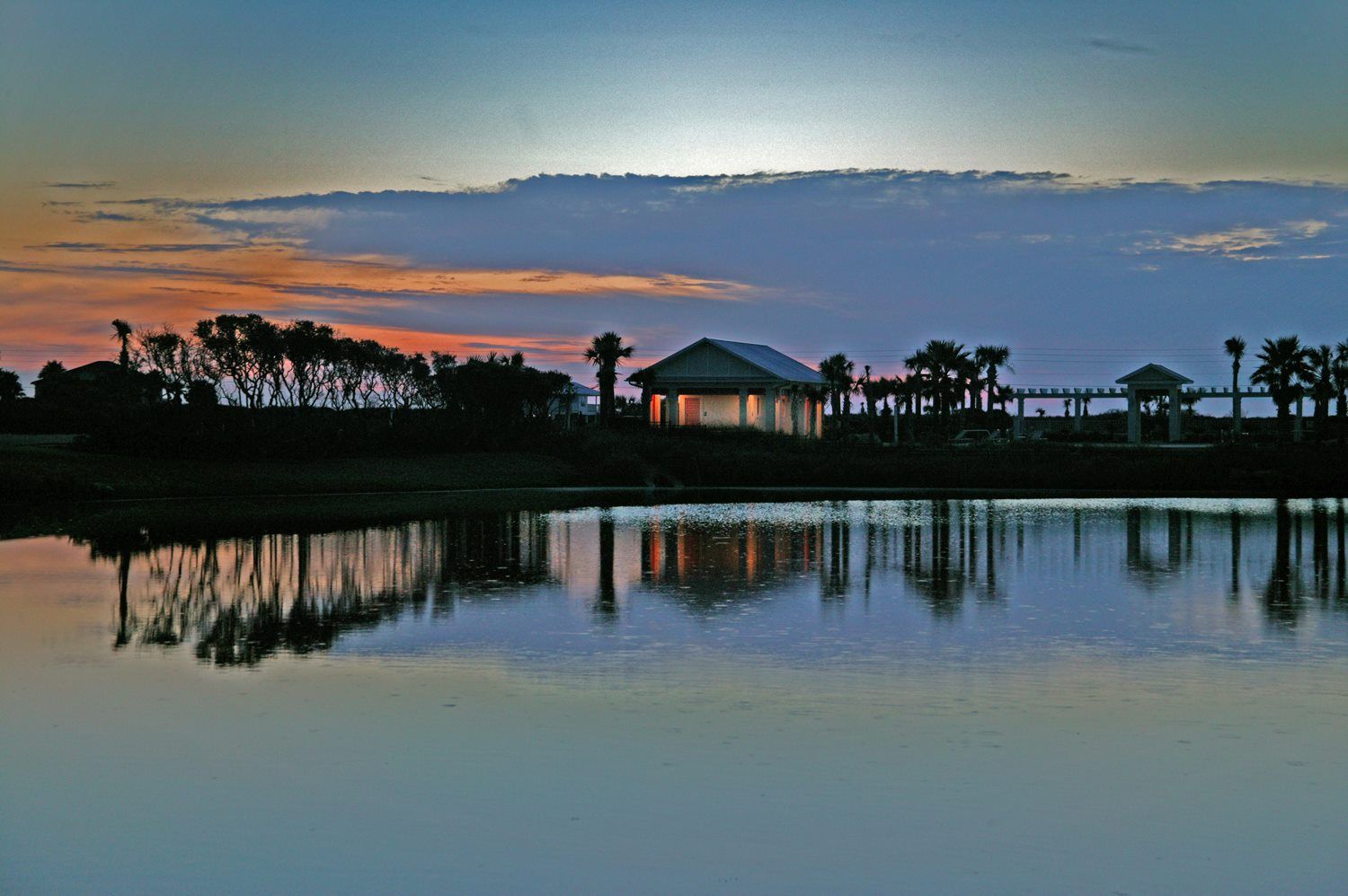 A sunset over a lake with a pavilion in the foreground and trees in the background.