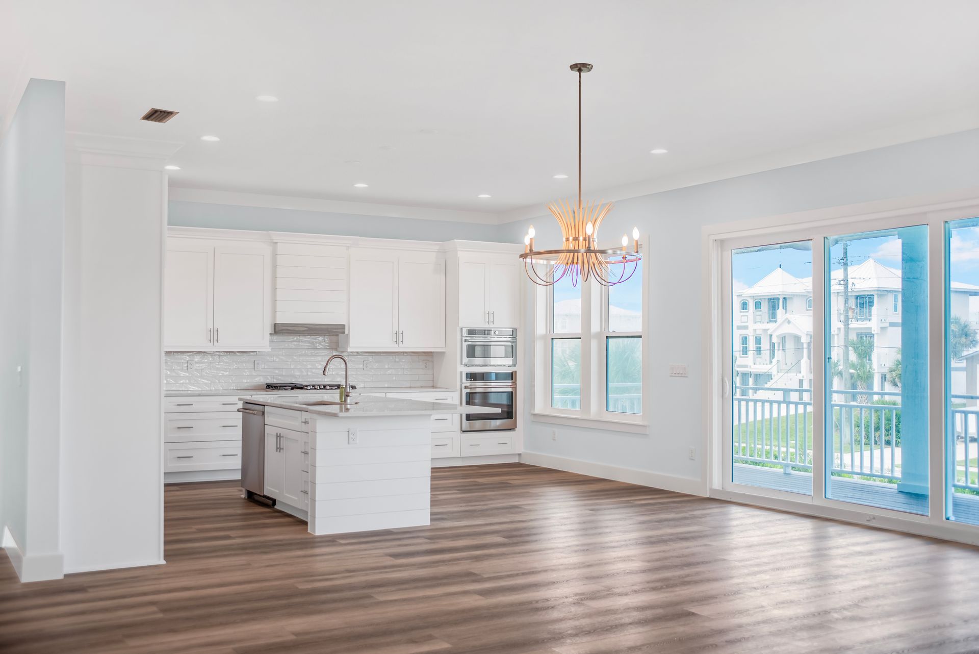 An empty kitchen with white cabinets , hardwood floors , sliding glass doors and a chandelier.