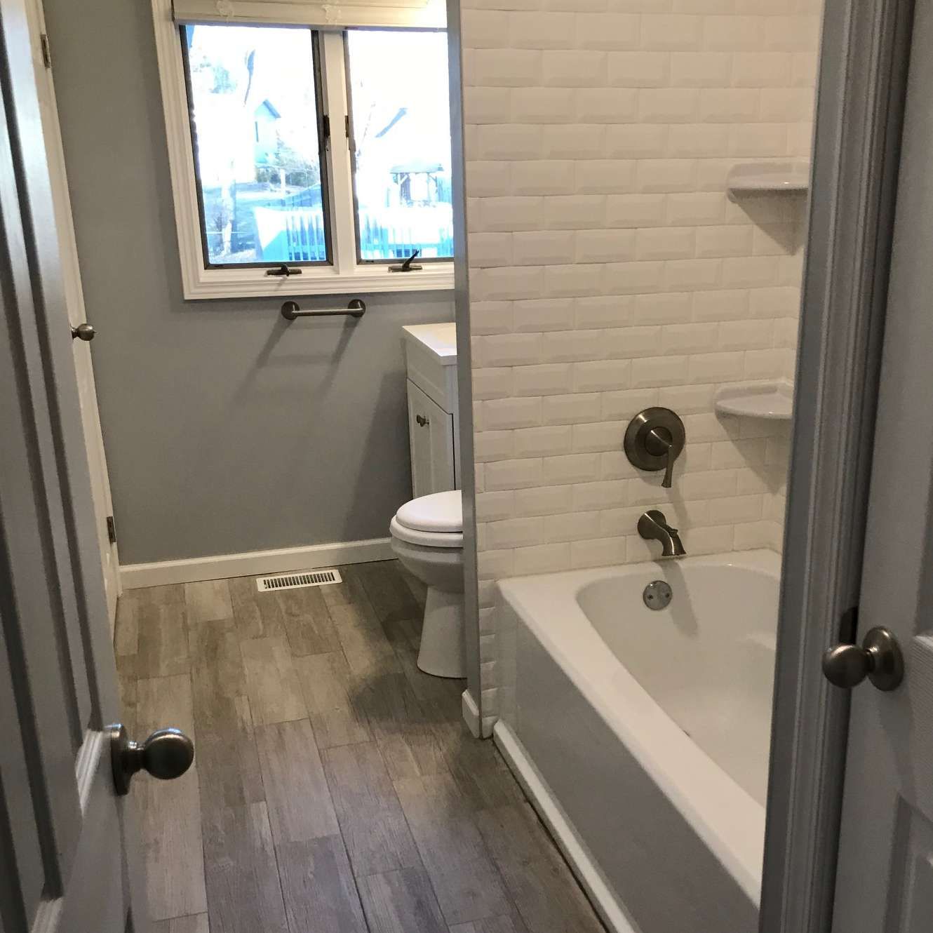 Bathroom with white tile, a bathtub, a toilet, a window, and wood-look flooring.