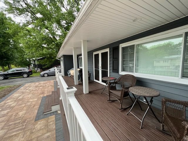 A house with a porch. Dark blue siding, brown deck, white railing, and outdoor furniture. 