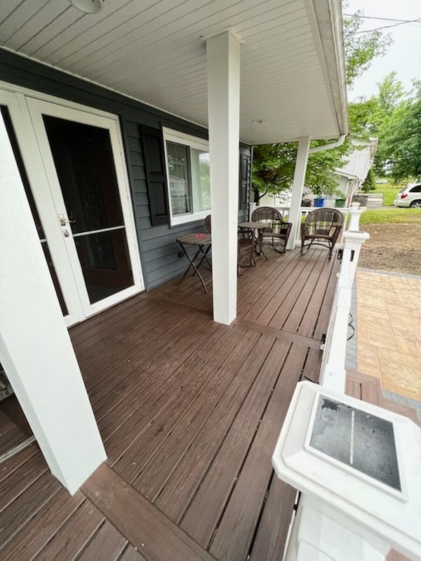 Porch with brown wooden deck, white columns, and dark siding; small table, chairs.