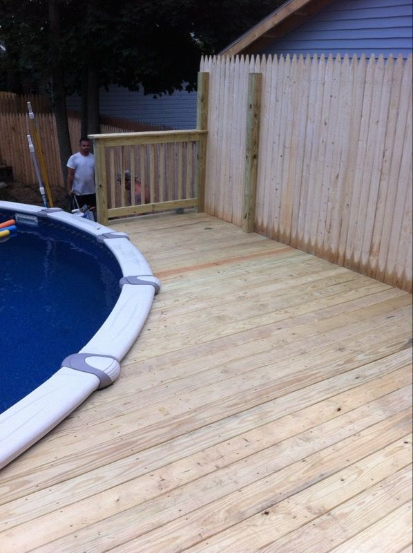 A wooden deck surrounds a pool with a fence and a person standing near the pool.