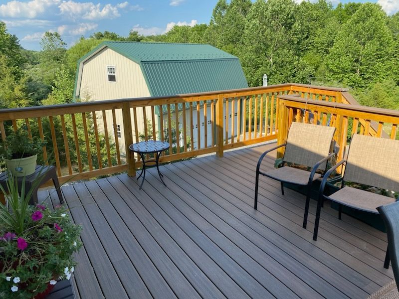 Wooden deck with two chairs, small table, and greenery overlooking a green-roofed building and trees.
