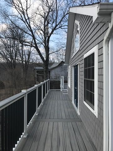 Gray deck with black railing and shingled house in a wooded area.