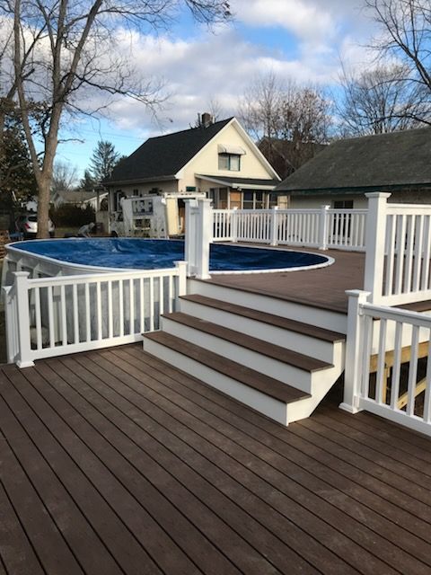 Brown deck with white railing surrounding an above-ground pool; house in background.