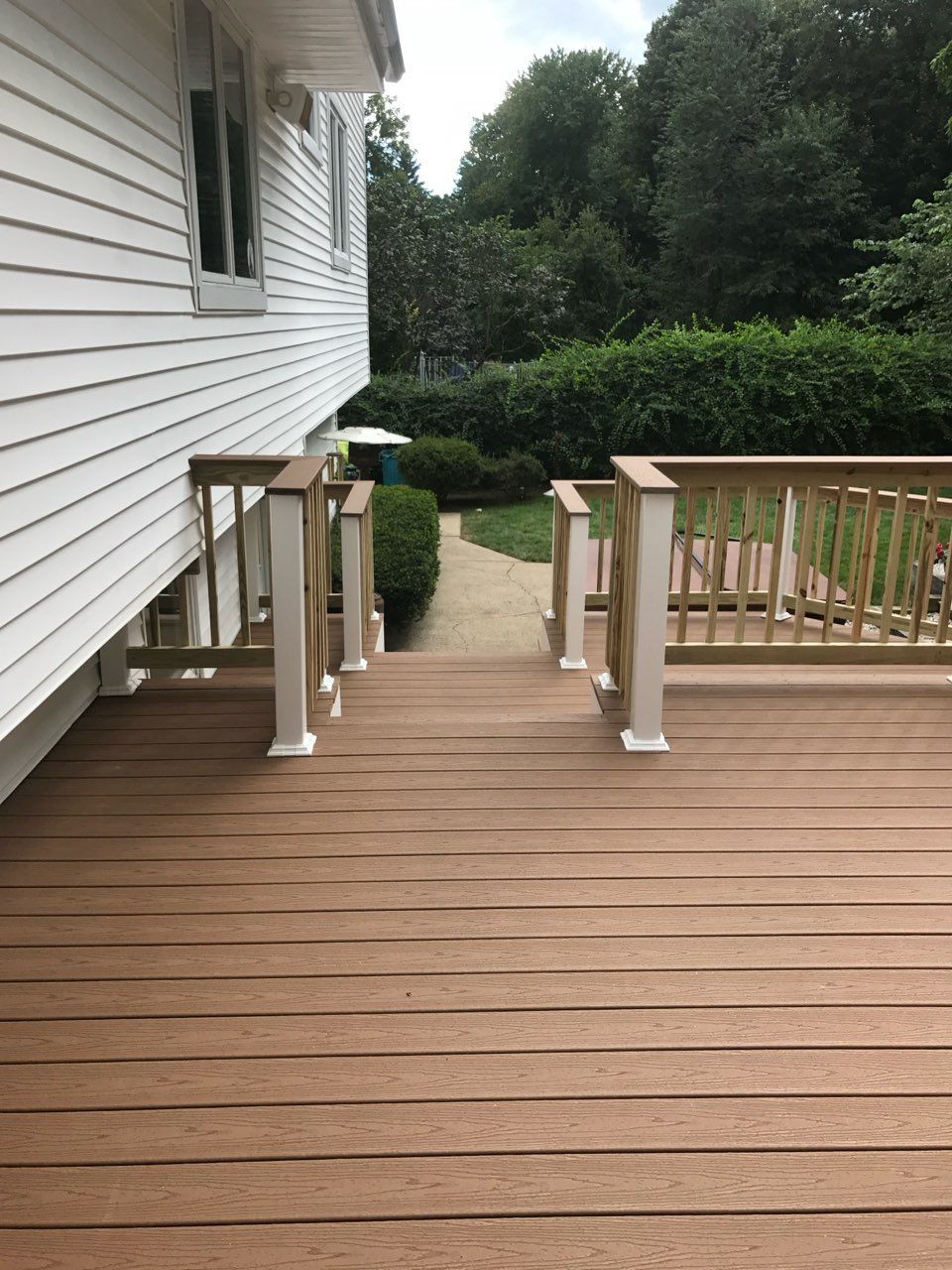 Brown wooden deck with steps leading to a path in a green yard, next to a white house with a deck railing.