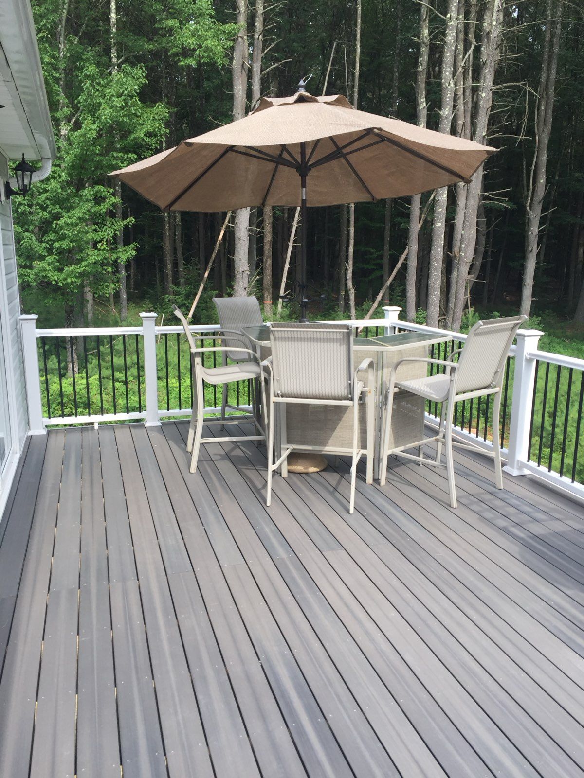 Outdoor deck with table and chairs under a tan umbrella; trees in the background.
