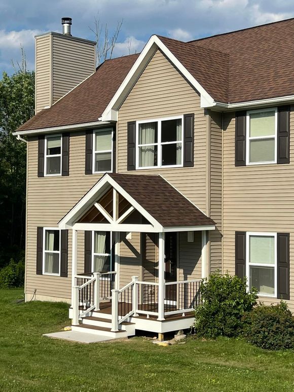 Tan two-story house with brown roof and porch, dark shutters, chimney, on a grassy lawn.