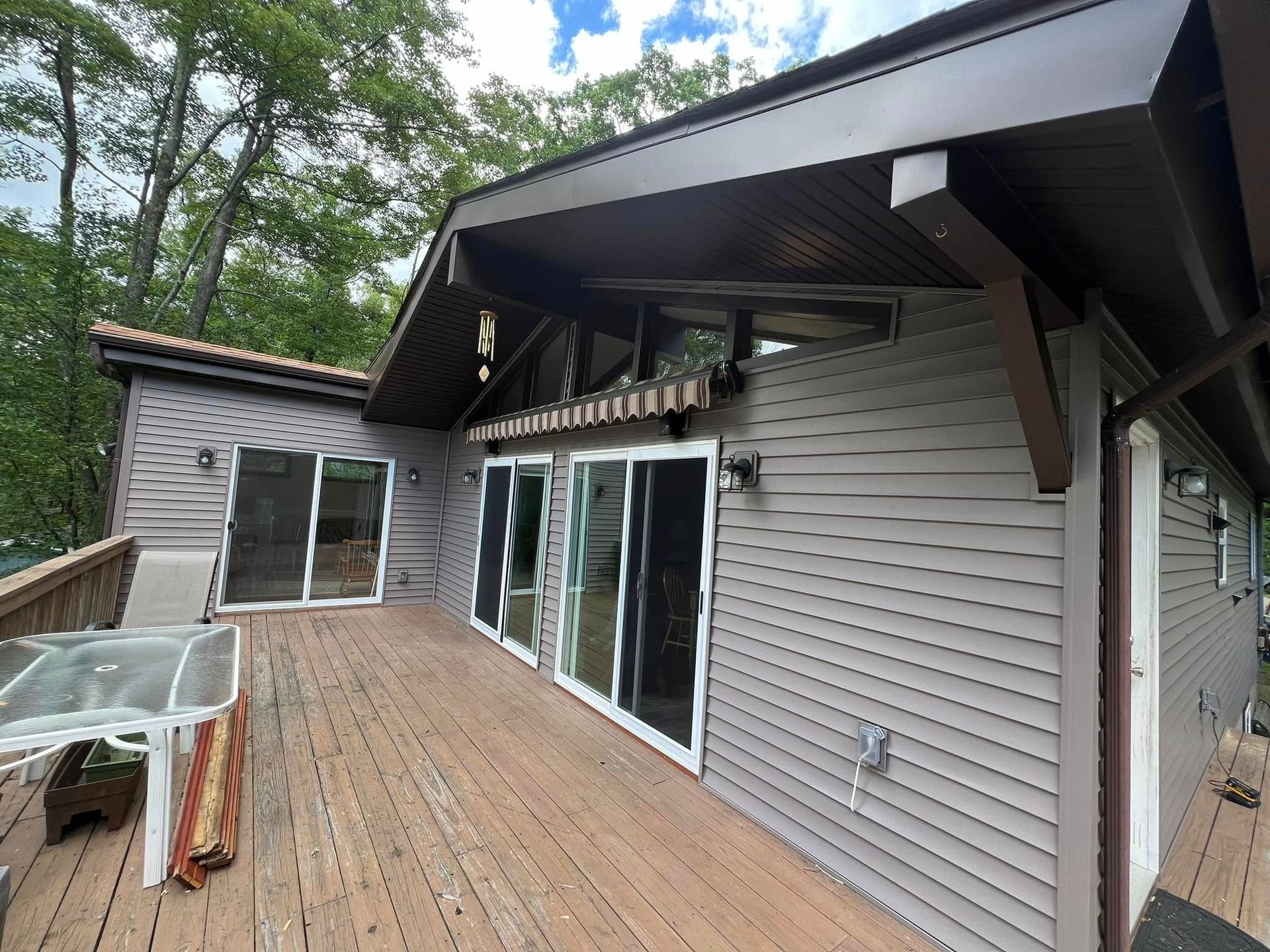 Brown and gray house with a wooden deck, glass sliding doors, and a tiled roof, trees in the background.