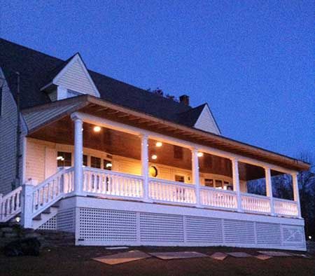 Lit porch with white railing and lattice, under a house at dusk.