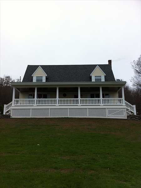 Yellow house with white porch and railings, two dormers, and a chimney on a grassy hill under an overcast sky.