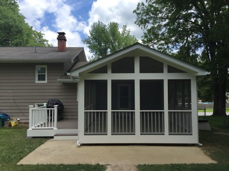 White screened-in porch addition with a gray roof next to a light brown house on a sunny day.