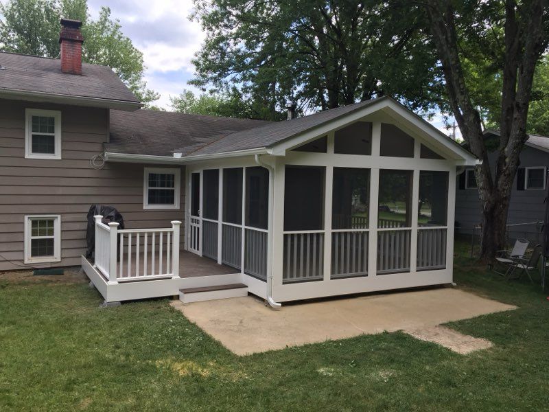 A screened-in porch addition with white railing and a gray roof, attached to a light brown house on a lawn.
