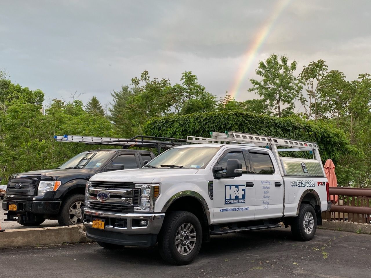 Two service trucks parked, one black and one white, with ladders on top, under a rainbow.