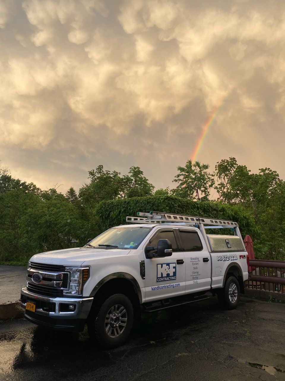 White truck with company logo parked under a rainbow, trees in the background, overcast sky.