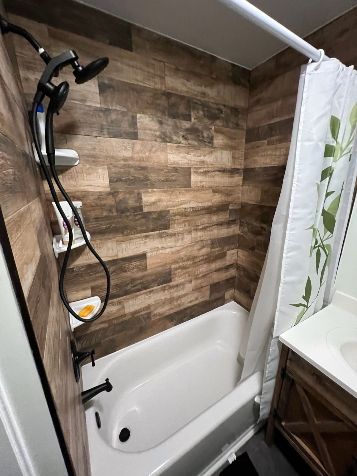 A small bathroom with a white bathtub and wood-look tile walls. A shower curtain with green leaves is pulled to the side.
