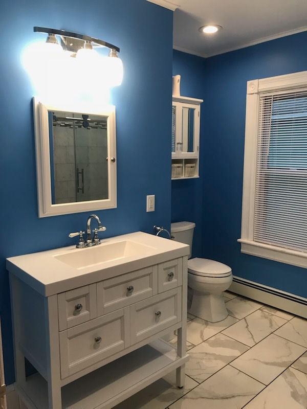 Blue bathroom with white vanity, toilet, and marble-look floor. White-framed mirror and window.