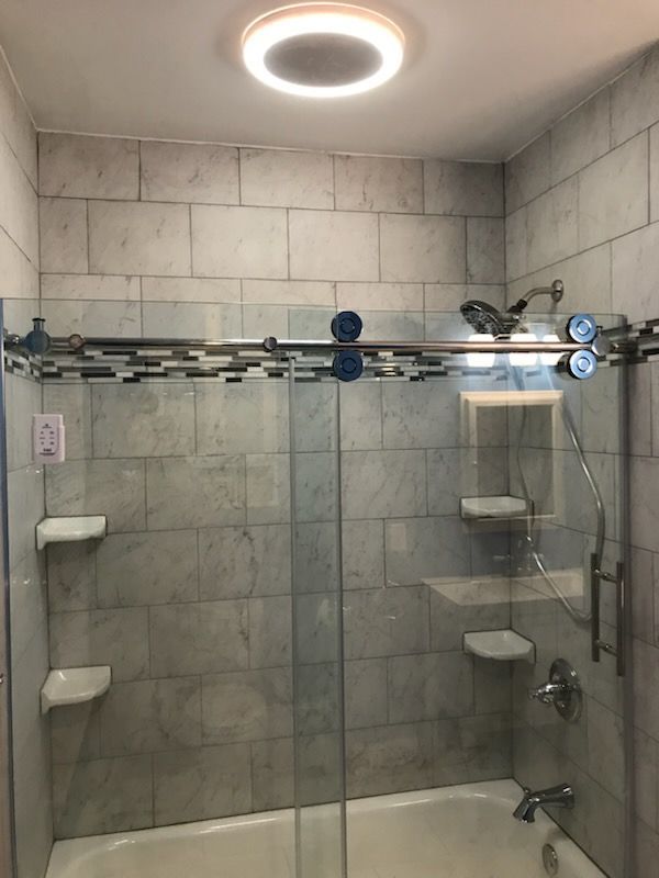 Shower stall with gray tiled walls, glass door, and overhead light. Soap shelves on the wall.