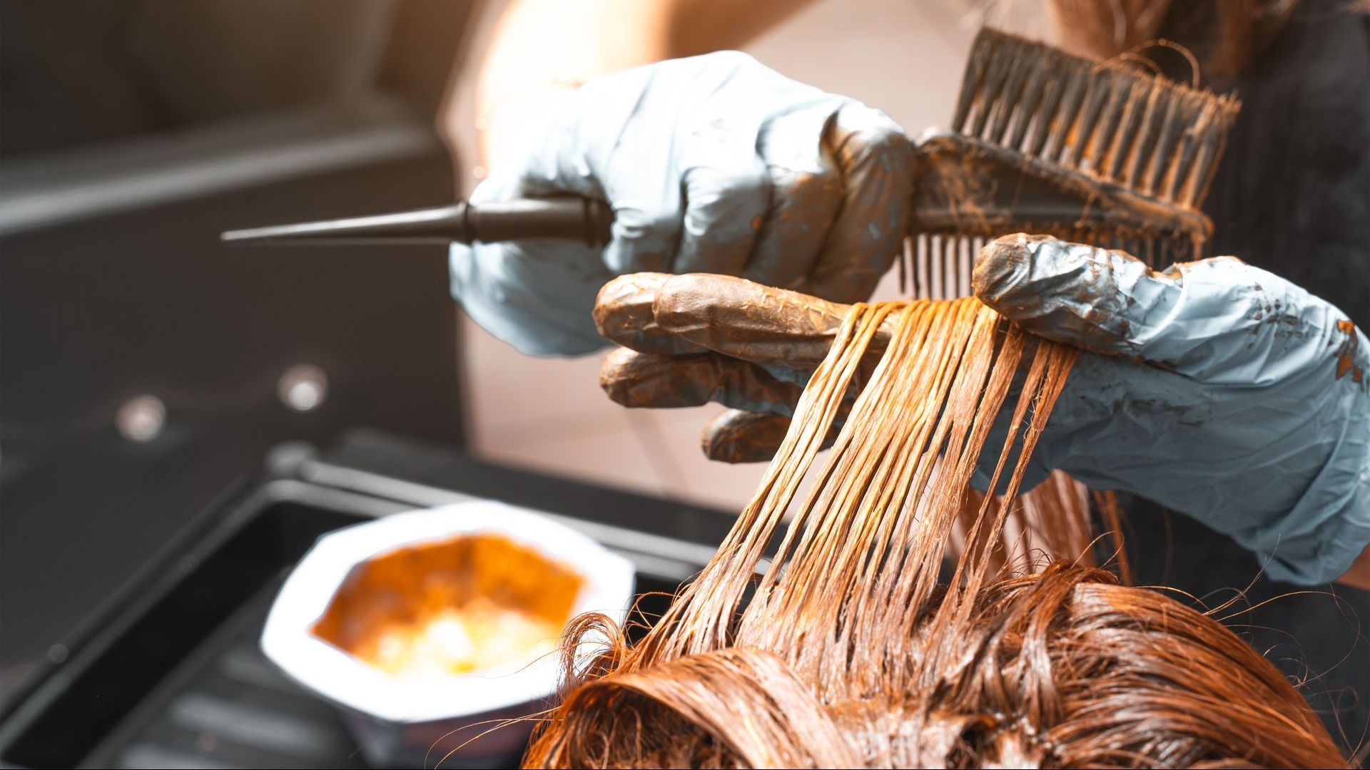 Hands in blue gloves applying hair dye with a brush and comb to red hair in a salon setting.