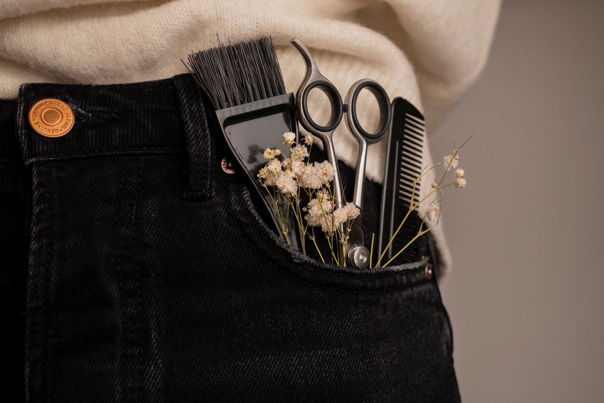 Hairdressing tools in a pocket: comb, shears, brush, and flowers in a black jeans pocket with a beige sweater.