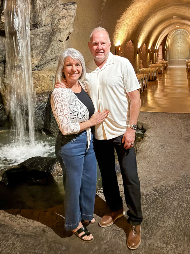 Jeff and Jeanine smiling, posing near a waterfall in a wine cellar with arched ceiling and barrels.