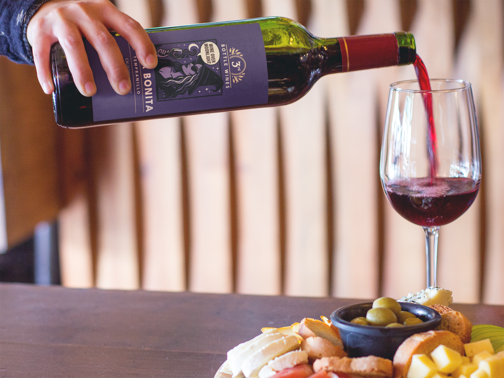 Hand pouring red wine from a bottle into a glass, charcuterie board in front.