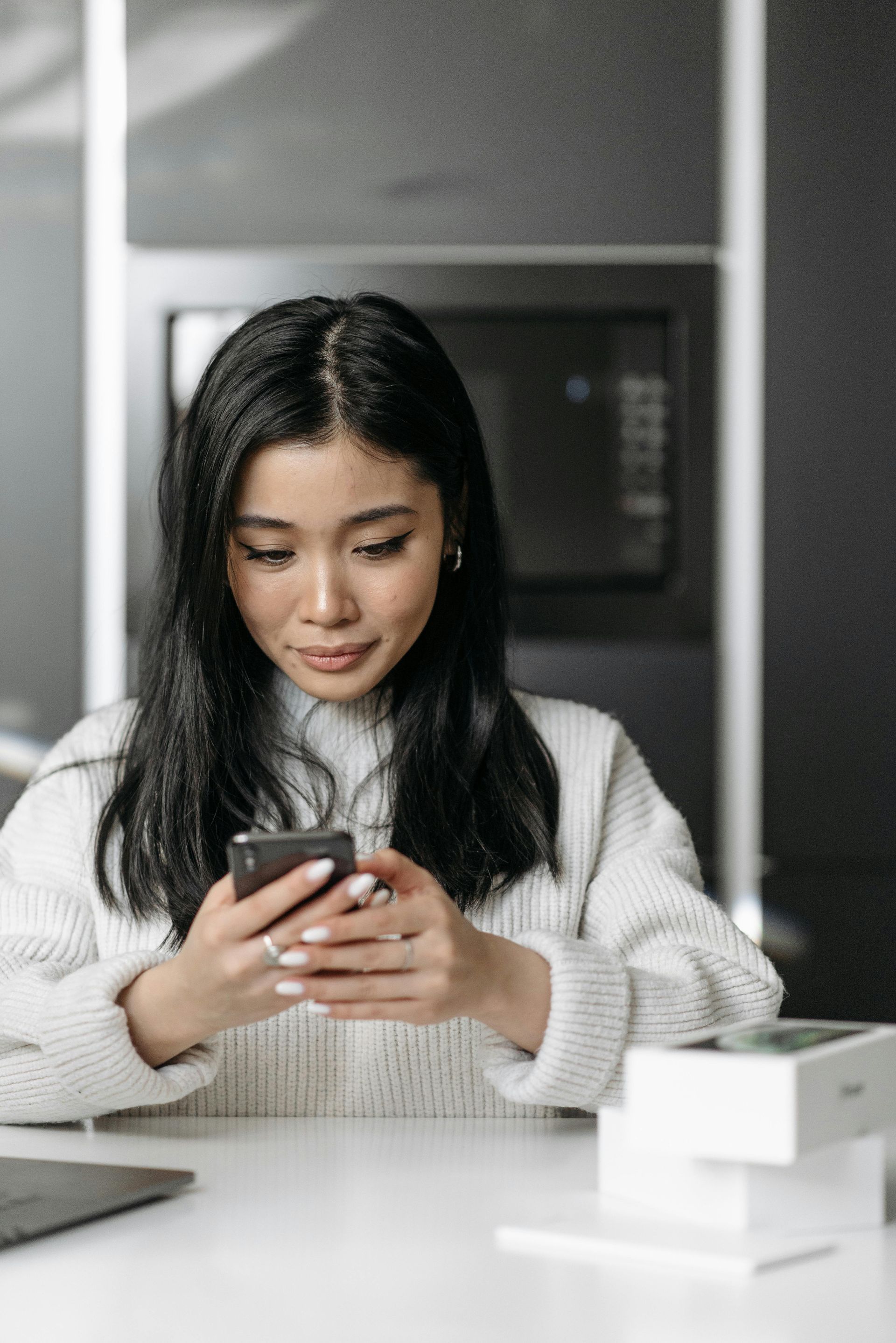 Woman with black hair, looking at phone, wearing a white sweater, in front of a white table.