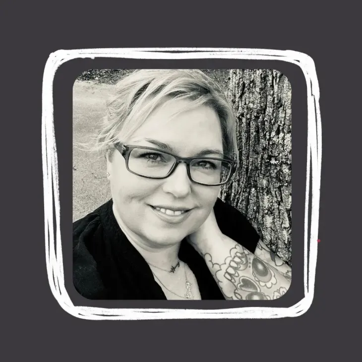 Woman with glasses and tattoo smiles at the camera, resting her head on her hand; black and white.