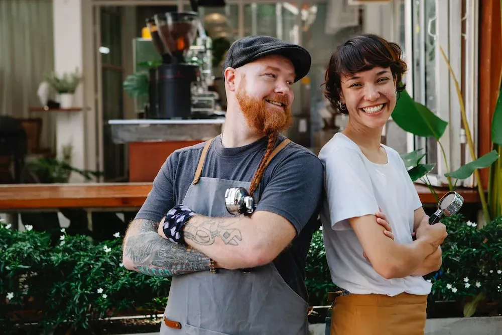 Couple with arms crossed, smiling.  They are wearing aprons and standing in front of a cafe.
Img by Tim Douglas - Pexels