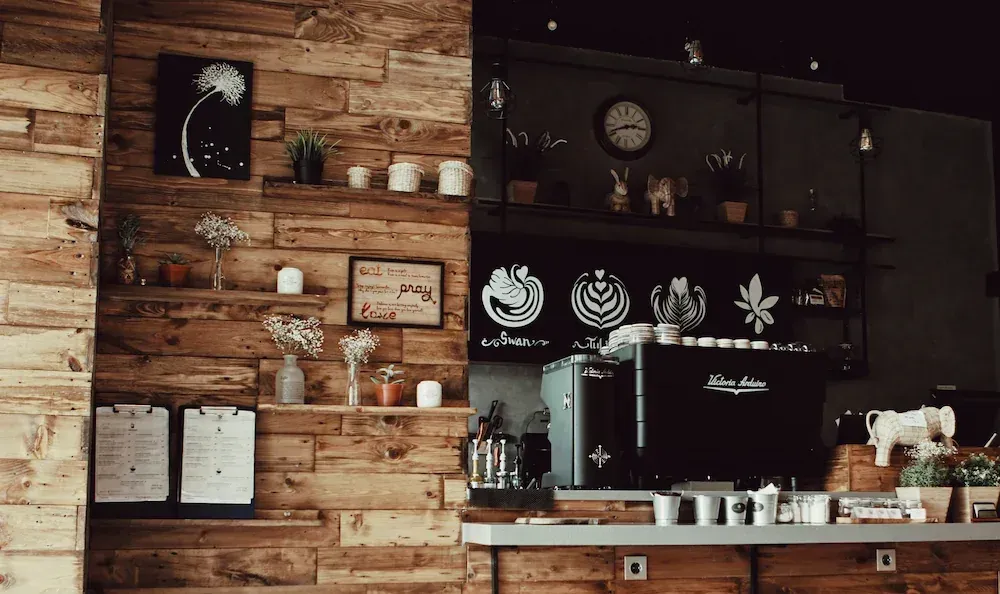Coffee shop interior: wooden wall with shelves, coffee machine, clock, decor, and menu boards.
Img by A. P. Gunawan - Pexels