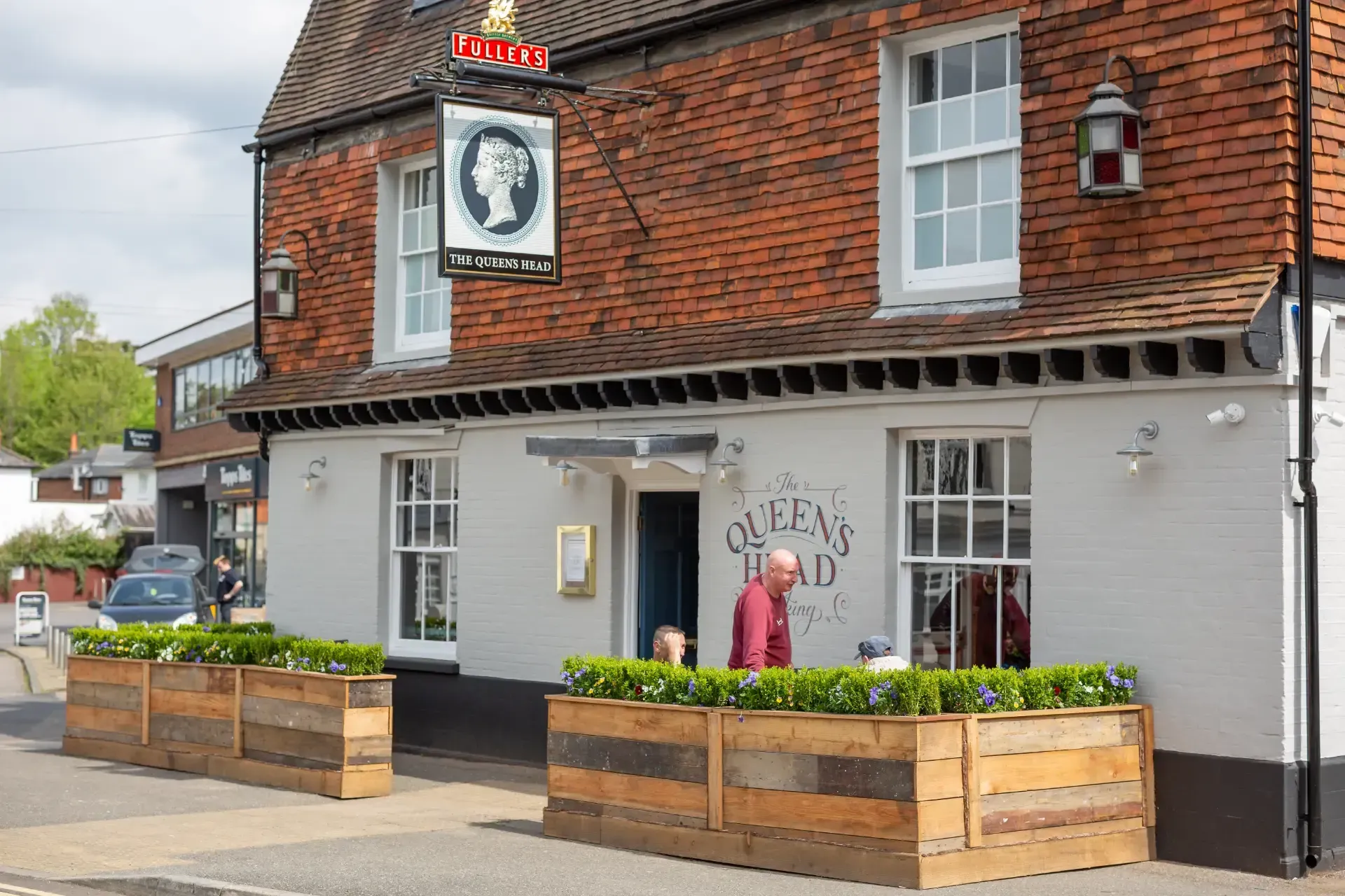 Exterior of "Queen's Head" pub with sign. White building with wooden planters. Person standing in the doorway.