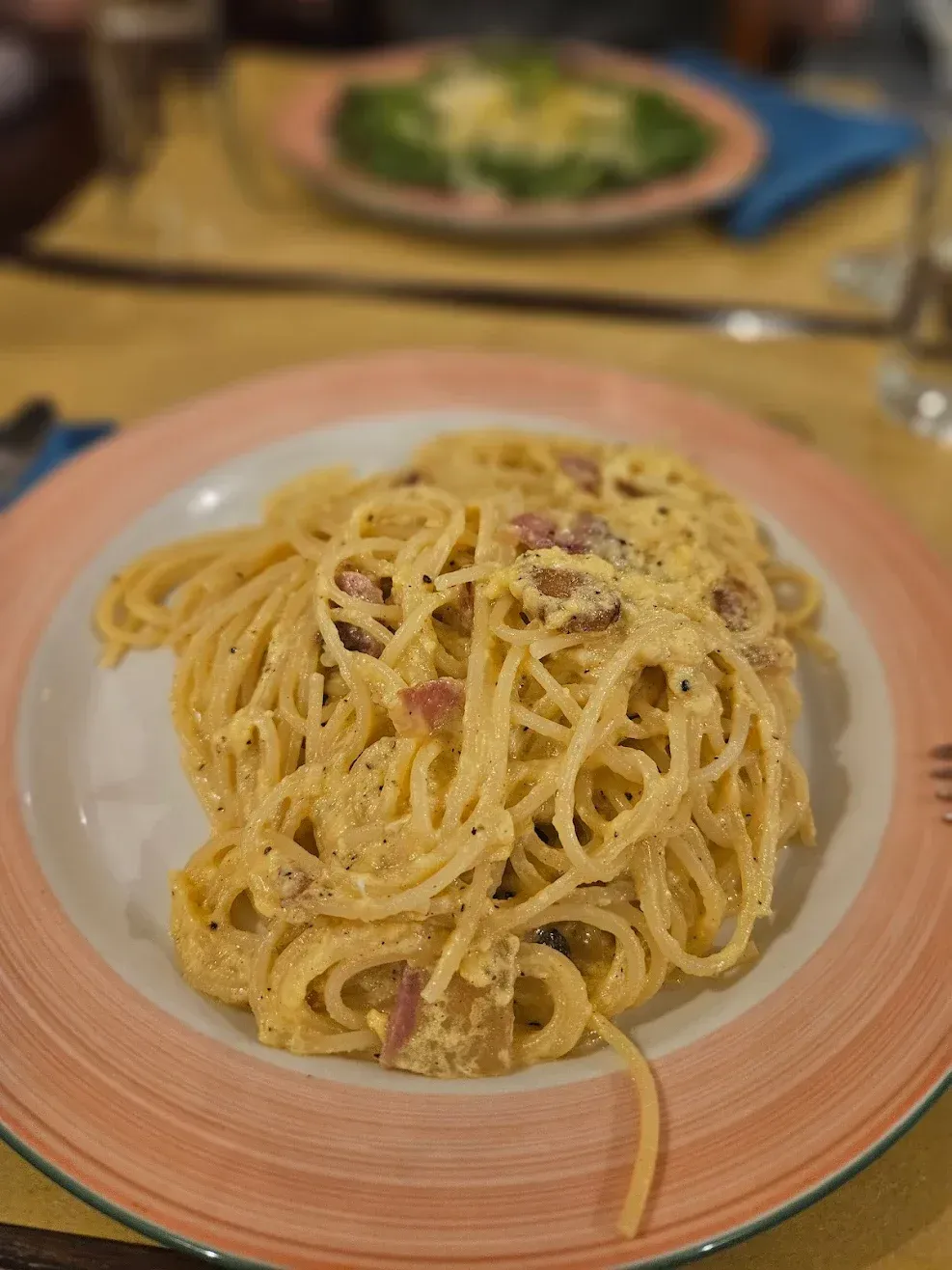 Un piatto di spaghetti alla carbonara cremosi su un piatto dal bordo rosa, con un piatto di pasta sfocato sullo sfondo.