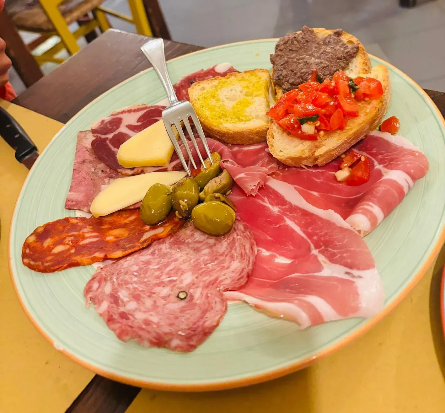 Piatto di salumi con affettati, olive, formaggio e bruschette su un piatto verde.