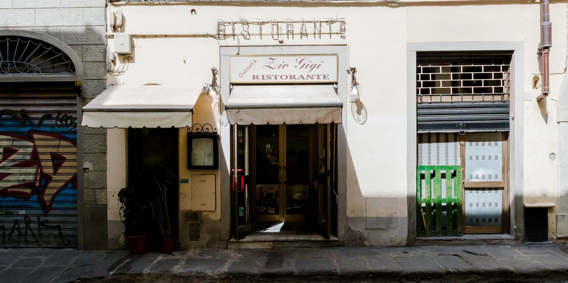 Negozio chiuso con facciata bianca, tenda da sole e porta adiacente con sbarre e bidoni della spazzatura verdi.