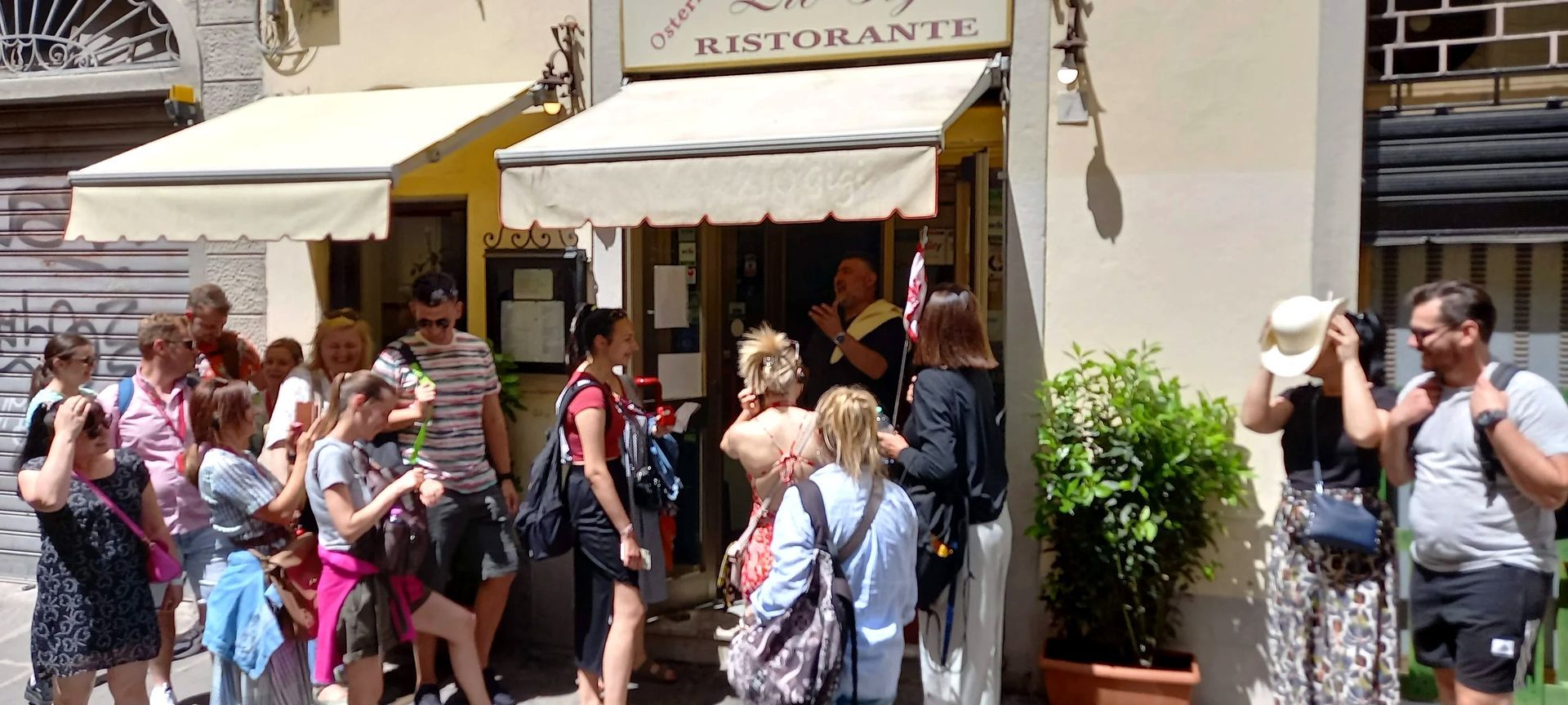 Una folla si è radunata davanti all'ingresso di un ristorante in una strada soleggiata, e alcune persone hanno scattato fotografie.