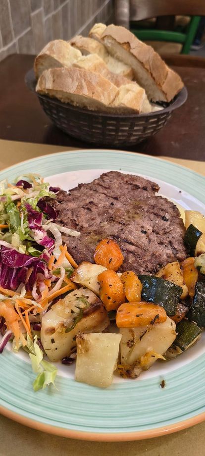 Piatto di bistecca alla griglia con verdure arrosto e cestino di pane sullo sfondo.