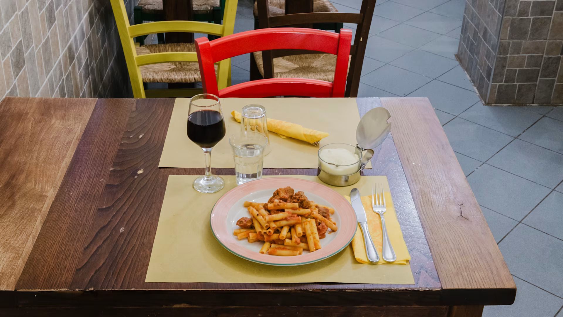 Un piccolo tavolo da ristorante apparecchiato con patatine fritte, bicchieri, posate e una sedia rossa sullo sfondo.