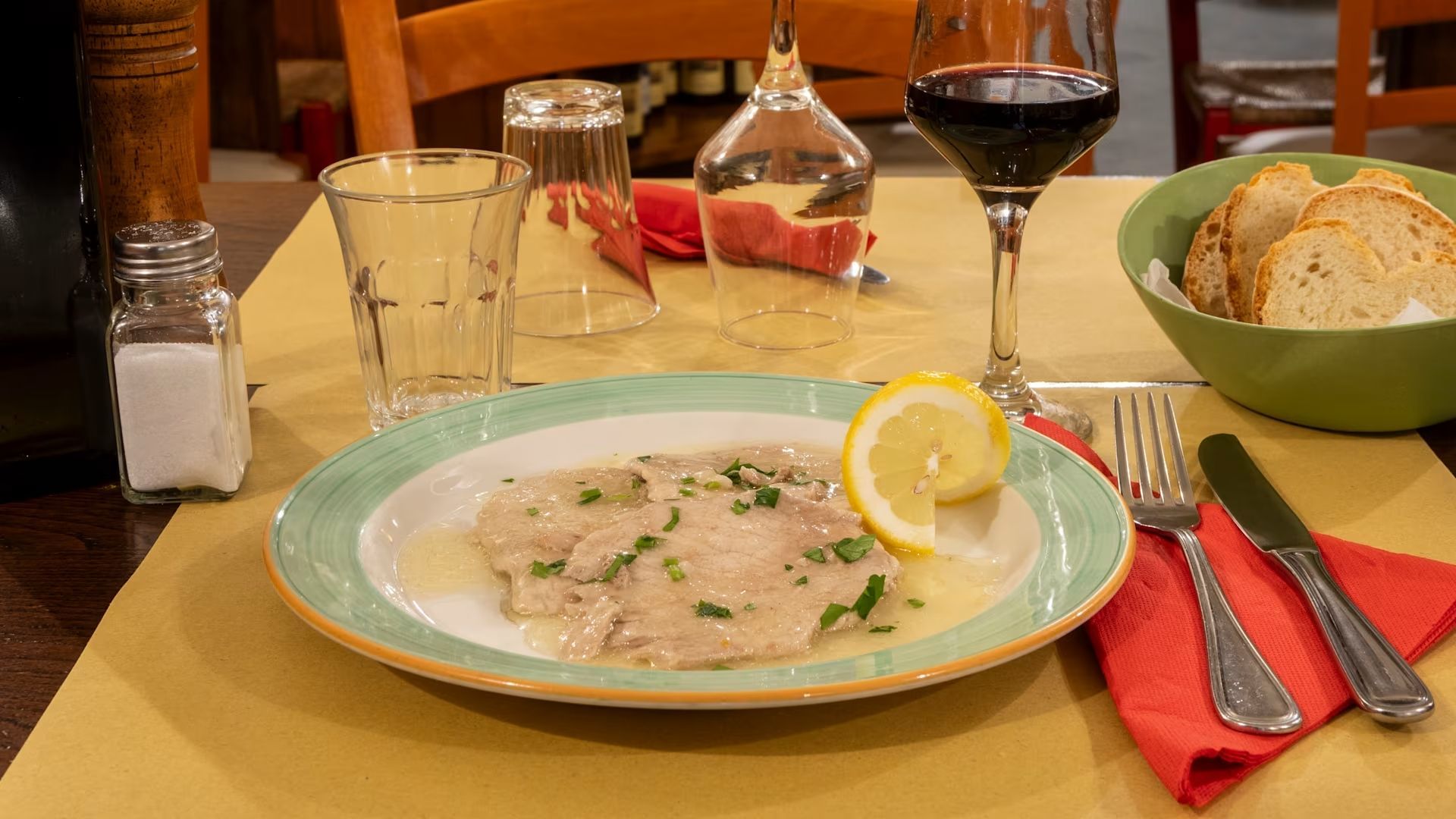 Tavola apparecchiata con pasta cremosa, guarnizione di limone, ciotola di pane e bicchieri di vino rosso su un tavolo di ristorante.