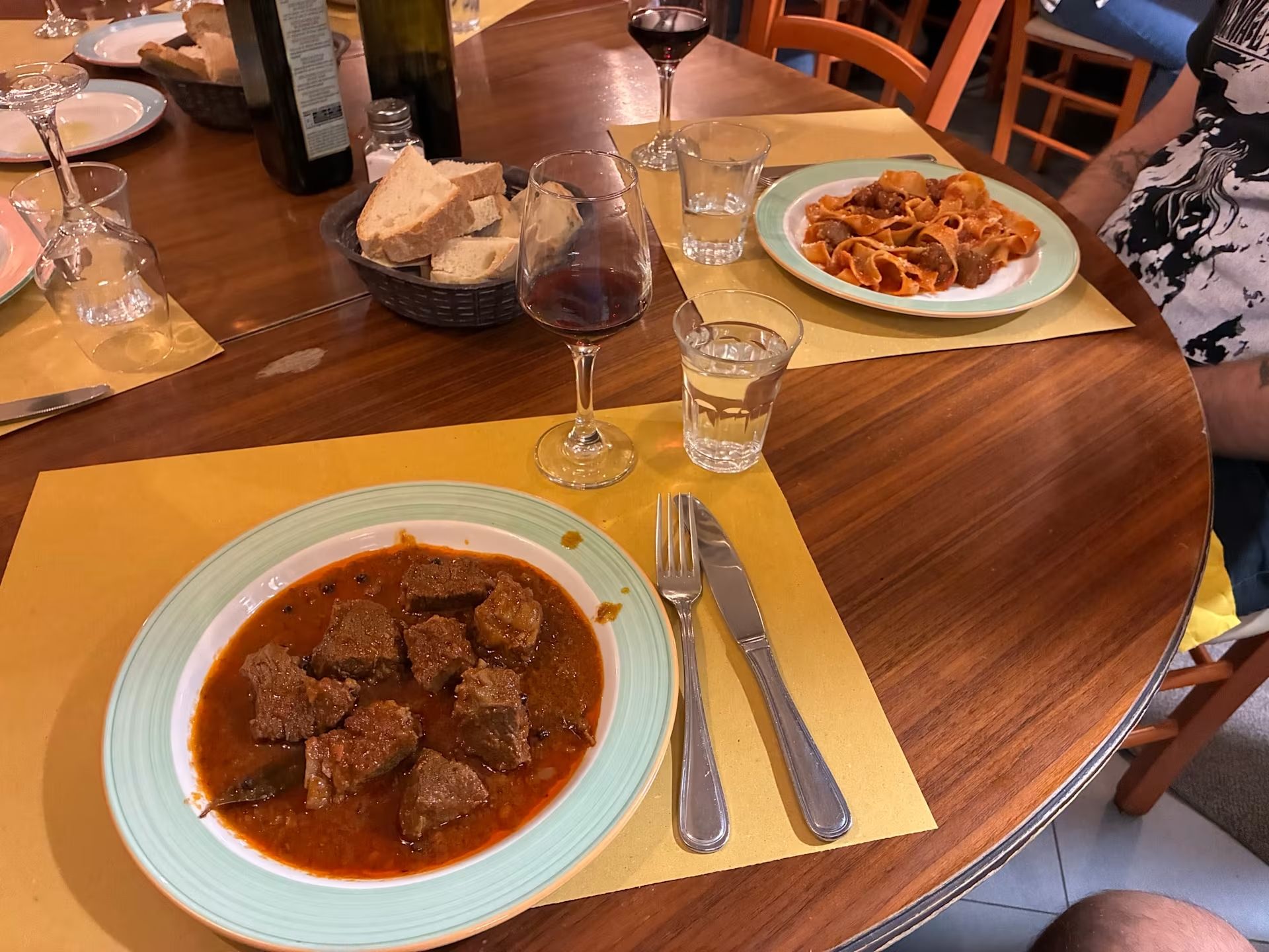 Tavolo da ristorante con due piatti di pasta e carne, bicchieri di vino rosso e pane su un tavolo di legno.