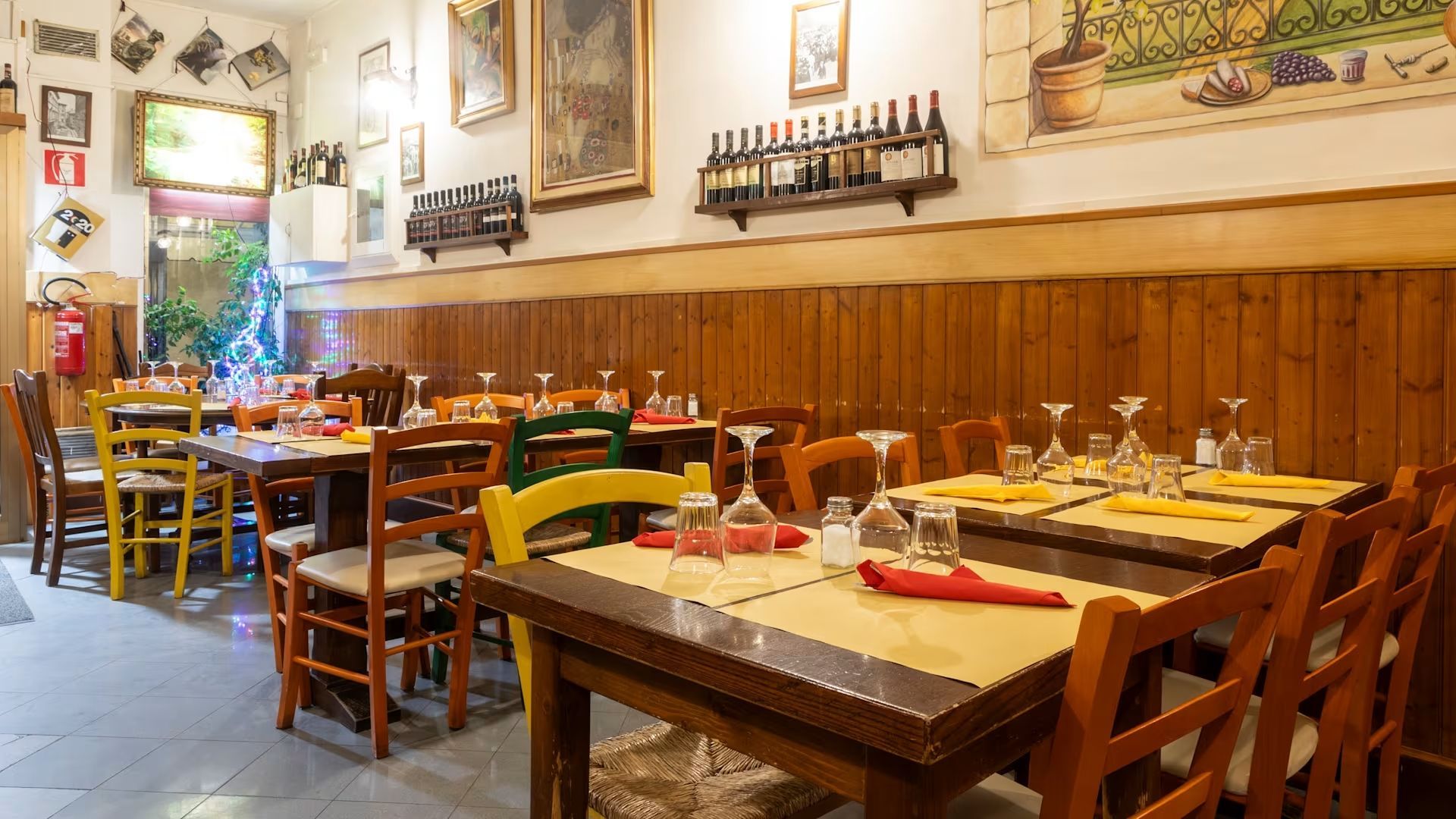 Accogliente sala da pranzo del ristorante con tavoli in legno, sedie rosse e bottiglie di vino appese al muro.